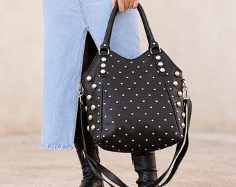 May include: A black handbag featuring a structured silhouette with two top handles and a detachable shoulder strap. The exterior is decorated with numerous silver-toned metal studs of varying sizes, arranged in a grid pattern across the front.