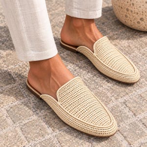 May include: A pair of beige woven slip-on mules featuring a textured, grid-like pattern across the upper. The shoes have a rounded toe, a low-profile silhouette, and a flat brown sole, paired with light-colored trousers that end above the ankle.