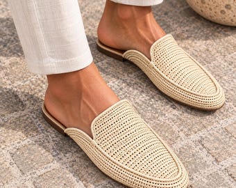 May include: A pair of beige woven slip-on mules featuring a textured, grid-like pattern across the upper. The shoes have a rounded toe, a low-profile silhouette, and a flat brown sole, paired with light-colored trousers that end above the ankle.