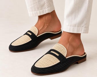 May include: A pair of backless loafers featuring a woven texture with a two-tone design. The shoes have a black outer border and a beige center panel on the vamp, finished with a low brown heel and a rounded toe shape for a slip-on style.