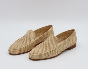 May include: A pair of beige woven loafers featuring a textured straw-like exterior, a rounded toe, and a low-profile silhouette. The shoes are finished with a smooth, dark brown leather sole and a small, flat heel, providing a clean, structured look.