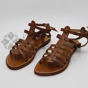 Może przedstawiać: Para brązowych skórzanych sandałów gladiatorów z wieloma paskami i klamrami. Sandały mają pleciony wzór i płaską podeszwę.