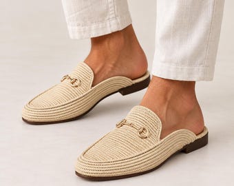 May include: A pair of beige woven slip-on mules featuring a gold-toned metal horsebit detail across the vamp. The shoes have a closed-toe design with a textured straw-like material and a low, flat dark brown heel, worn with white trousers.