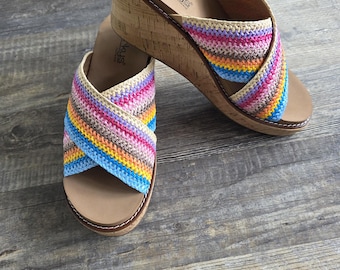 May include: A pair of Corkys wedge sandals with a multi-colored woven strap design. The sandals have a cork wedge heel and a tan footbed. The straps feature a rainbow of colors including pink, blue, yellow, and orange. The sandals are displayed on a wooden surface.