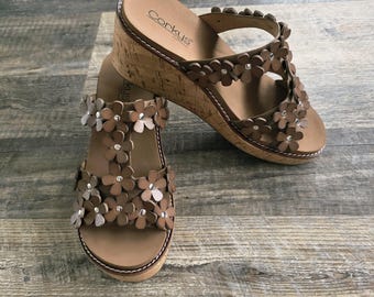 May include: Brown wedge sandals with floral embellishments. The sandals feature a cork sole and a strap design adorned with flower-shaped details. The brand name "Corkys" is visible on the side.