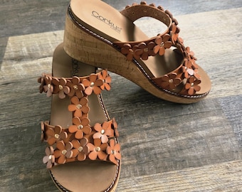 May include: A pair of brown leather wedge sandals with floral accents. The sandals feature a cork wedge heel and a strap adorned with small, flower-shaped cutouts. The footbed is a light brown color, and the sole has white stitching. The brand name "Corkys" is visible.