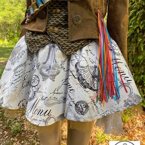 May include: A white skirt with a vintage-inspired print featuring a menu, a teapot, and a clock. The skirt has a gold tulle overlay and is paired with a brown jacket with gold accents and a colorful ribbon.