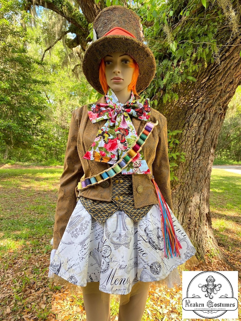 May include: A brown and gold Mad Hatter costume with a tall brown top hat, a brown jacket with a gold vest, a white and black patterned skirt, and a colorful bow tie. The costume is worn by a mannequin with orange hair.