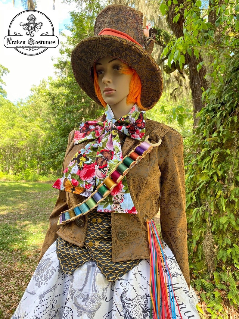 May include: A brown and gold brocade jacket with a colorful butterfly print bow tie and a spool of thread belt. The jacket has a gold and black vest underneath. The person is wearing a white skirt with a black and white print and a tall brown top hat with a red band.