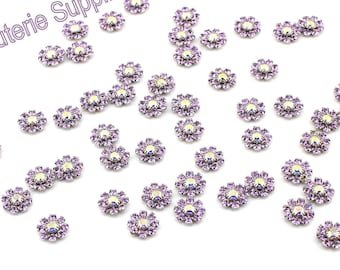 May include: A scattering of small, purple flower-shaped rhinestones with iridescent centers. The rhinestones are scattered on a white background.