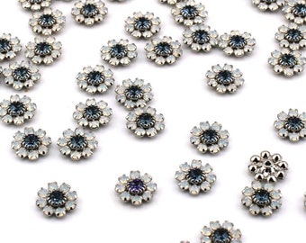 May include: A scattering of silver-toned flower-shaped embellishments with iridescent blue centers. The embellishments are scattered on a white surface.
