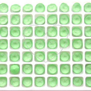 May include: A white tray containing 100 small, round, light green glass cabochons. The cabochons are arranged in a grid pattern.