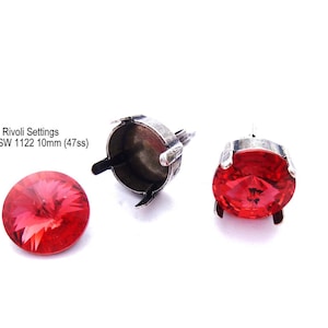 May include: Two silver stud settings for 10mm round rhinestones. The settings have four prongs and a flat back. One setting has a red rhinestone in place.