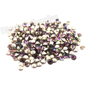 May include: A pile of iridescent rhinestones with a silver base. The rhinestones have a faceted cut, reflecting purple, green, and gold hues. The image is on a white background, with the text "Bijoux Supplies" in the upper left corner.
