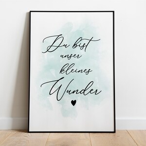 Op de afbeelding: Een witte poster met een zwarte lijst en een lichtblauwe aquarel achtergrond. De tekst op de poster luidt "Du bist unser kleines Wunder" met een klein zwart hartje onder de tekst.