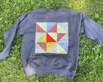 May include: A dark grey sweatshirt with a patchwork design on the front. The patchwork is made up of different coloured fabrics, including red, yellow, blue, and green. The sweatshirt has a crew neck and long sleeves.