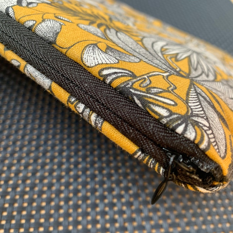 Pu&ograve; includere: Primo piano di una pochette con cerniera con un motivo floreale giallo senape. Il design presenta elementi botanici bianchi e grigi. La cerniera &egrave; nera e la pochette sembra essere di tessuto.