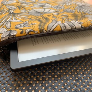 Pu&ograve; includere: Una custodia per e-reader con stampa floreale giallo senape e nera con cerniera nera. La custodia &egrave; parzialmente aperta, rivelando un e-reader nero con testo sullo schermo. La custodia &egrave; su una superficie tessuta.