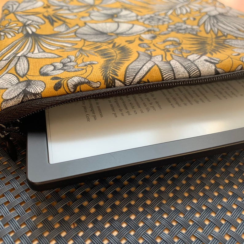 Pu&ograve; includere: Una custodia per e-reader con stampa floreale giallo senape e nera con cerniera nera. La custodia &egrave; parzialmente aperta, rivelando un e-reader nero con testo sullo schermo. La custodia &egrave; su una superficie tessuta.
