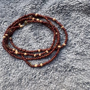 May include: A brown and beige beaded necklace with a gold clasp. The necklace is made of multiple strands of beads and is lying on a grey fluffy surface.