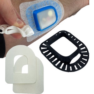 May include: A white medical adhesive bandage with a blue and white plastic container attached to it. The container has a clear lid and is designed to hold small items. There are two white foam pads with cutouts in the shape of a D. A black plastic ring with a square cutout is also shown.