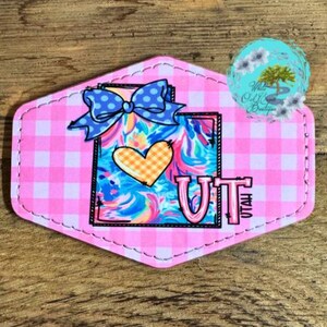 May include: A pink and white gingham background with a blue and white bow on top of a blue, pink, and yellow swirl design. The design is shaped like the state of Utah with a yellow heart and the letters "UT" in white. The words "UTAH" are written vertically in white on the right side of the design.