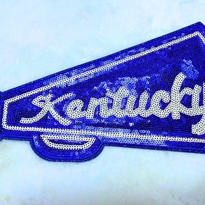 May include: A megaphone-shaped patch covered in royal blue sequins with a white sequined border and the word Kentucky written in white script across the center. The design features a small white sequined detail on the handle area.