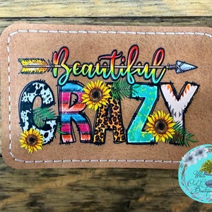 May include: A rectangular patch with the words "Beautiful Crazy" in colorful, stylized lettering. The design includes sunflowers, an arrow, and a cow print. The patch is on a tan background with white stitching.