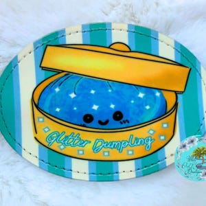 May include: An oval-shaped patch featuring a cartoon glitter dumpling in a yellow container with a lid. The dumpling is blue with white star accents and a smiling face. The text "Glitter Dumpling" is written in teal script. The background has teal and white stripes.