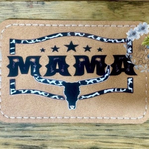 May include: A tan rectangular patch with the word "MAMA" in bold black letters. The design includes a longhorn skull, stars, and a border with a black and white cow print pattern. The patch is stitched with white thread.
