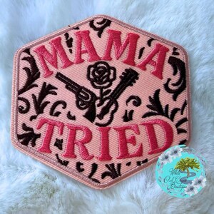 May include: A hexagonal embroidered patch with a light pink background and a dark brown filigree pattern. The text MAMA TRIED is stitched in bright pink thread. Centered between the words is an embroidered rose and two crossed revolvers.