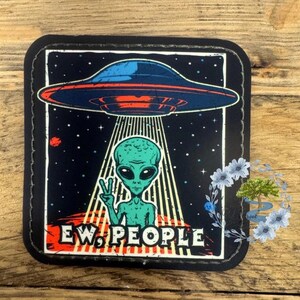 May include: A black and white patch with a green alien in a flying saucer. The alien is holding up two fingers in a peace sign. The text "EW. PEOPLE" is below the alien.