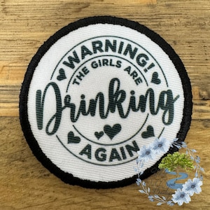 May include: A round, white patch with black trim features the text "WARNING! THE GIRLS ARE Drinking AGAIN" in a dark green script. The design includes heart shapes. The patch is on a wooden surface.