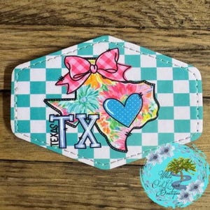 May include: A white and teal checkered background with a pink bow and a floral design of the state of Texas. The text "TEXAS" is written in blue and white letters. A blue heart with white polka dots is in the center of the design.