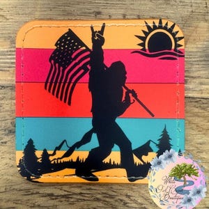May include: A colorful coaster with a silhouette of a Bigfoot holding an American flag and a sunset in the background.
