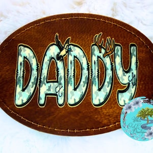 May include: Brown oval-shaped patch with the word "DADDY" in a camouflage pattern. The letters are outlined in black and decorated with a duck and antlers. The patch has a stitched border and a small logo in the bottom right corner.
