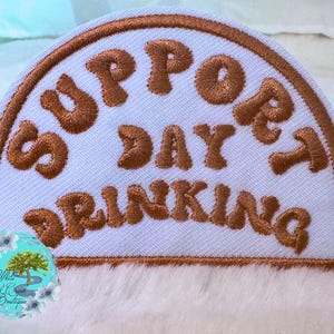 May include: A white, semi-circular patch with brown embroidered text that reads "SUPPORT DAY DRINKING". The patch has a brown border and is set against a soft, white background. A small, round logo with a tree and flowers is in the lower left corner.