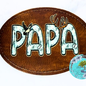 May include: A brown, oval-shaped patch with the word "PAPA" in large, light blue camouflage letters. A duck and deer antlers are above the letters. The patch has a stitched border and a small, round logo in the bottom right corner.