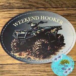 May include: Oval sticker with the text "WEEKEND HOOKER" above an ATV with camouflage detailing. The ATV is in mud, and the sticker has a stitched border. A floral logo is in the bottom right corner.