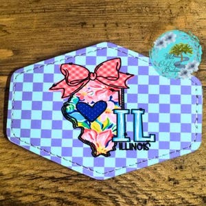 May include: A blue and purple checkered background with a floral outline of the state of Illinois. The outline has a blue heart in the center and a pink bow at the top. The text "IL ILLINOIS" is below the outline.