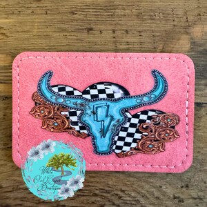 May include: Pink rectangular wallet with a turquoise bull skull design, a black and white checkered pattern, and copper-colored filigree accents. The wallet has a stitched border and a logo that says "White Oak Creek Boutique".