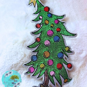 May include: A glittery green Christmas tree patch with colorful ornaments and a gold star on top. The tree has a silver outline and a brown trunk. The patch is on a white, fluffy surface.
