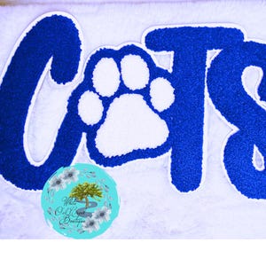 May include: A white, fluffy surface with the word "Cats" in blue, stylized lettering. The letter "O" is replaced with a white paw print. A turquoise circle with a floral design and text is in the lower left corner.