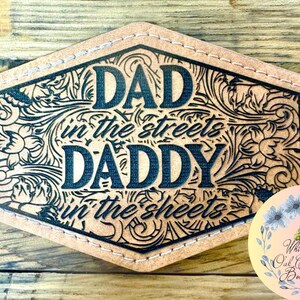 May include: A light brown leather patch with the words "DAD in the streets DADDY in the sheets" in black, surrounded by floral and scroll designs. The patch has a stitched border and a decorative floral logo in the bottom right corner.
