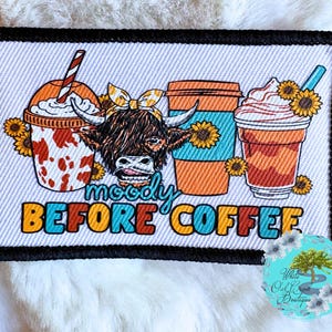 May include: A rectangular patch with a black border features a cartoon Highland cow with a yellow bow, surrounded by coffee drinks and sunflowers. The text "moody BEFORE COFFEE" is in colorful letters.