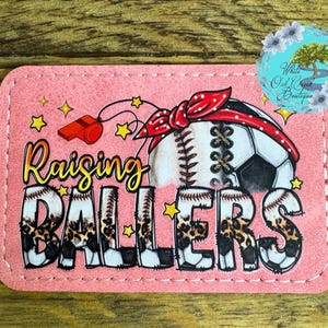 May include: Pink leatherette patch with a stitched border. The patch features a baseball and soccer ball with a red and white bandana on top. The text "Raising Ballers" is in black and white with a leopard print pattern.