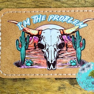 May include: A brown leather patch with a white cow skull and the text "I'm the problem" in blue and pink lettering. The skull is set against a desert landscape with cacti and a sunset.