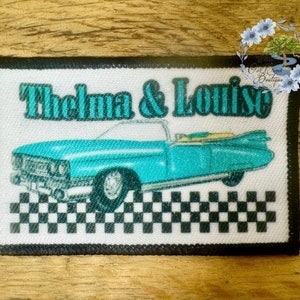 May include: A white fabric patch with black trim featuring a turquoise car, black and white checkered pattern, and the text "Thelma & Louise".
