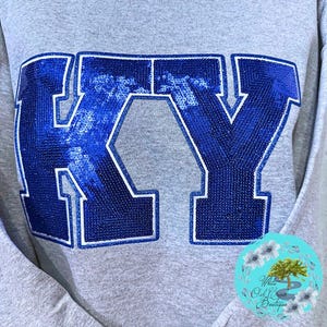 May include: A gray sweatshirt with the letters "KY" in blue sequins outlined in white. The sweatshirt has a crew neck and long sleeves. A circular logo with text is visible in the lower right corner.