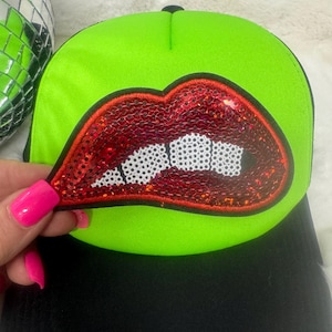 May include: A neon green baseball cap with a black brim. The cap features a sequined red lips design with white teeth. The cap is held by a hand with pink nail polish.